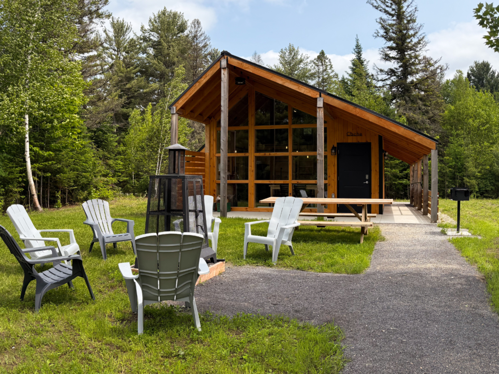 Chalet en bois de Chalets Lanaudière avec terrasse, table de pique-nique et fauteuils extérieurs, entouré d’arbres.
