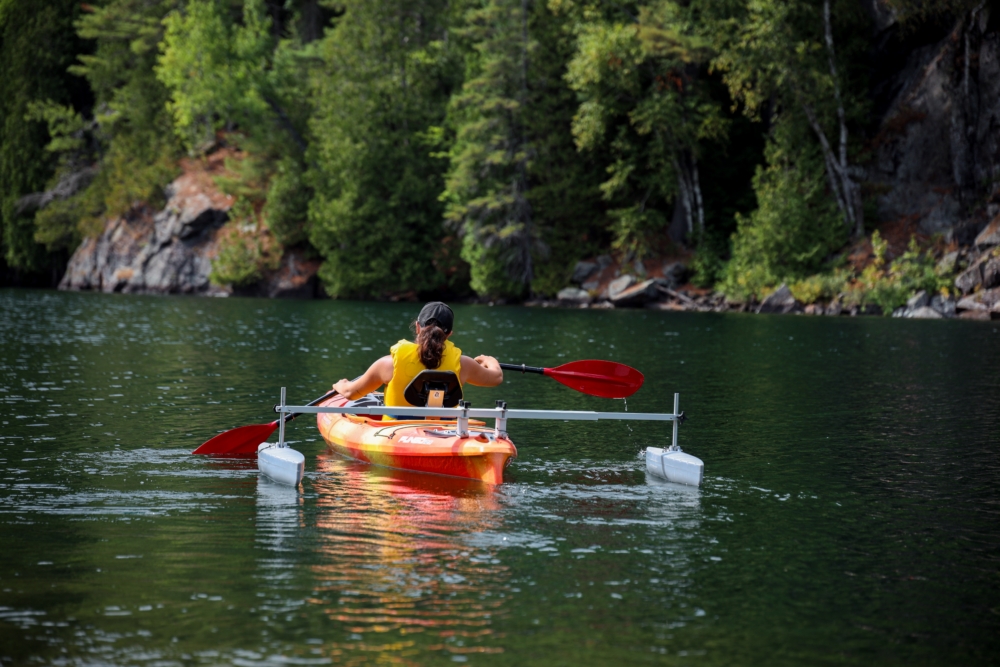 Personne en kayak orange sur un lac boisé, près d’un quai flottant, avec une pagaie rouge.