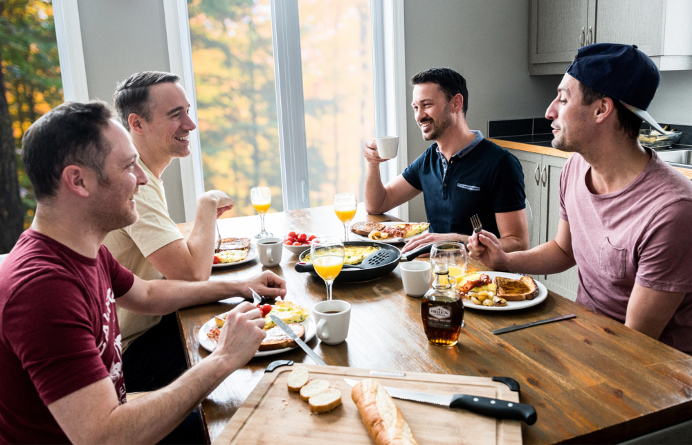 Quatre personnes partagent un repas autour d’une table en bois, avec assiettes, boissons et pain, dans une cuisine lumineuse près des fenêtres.