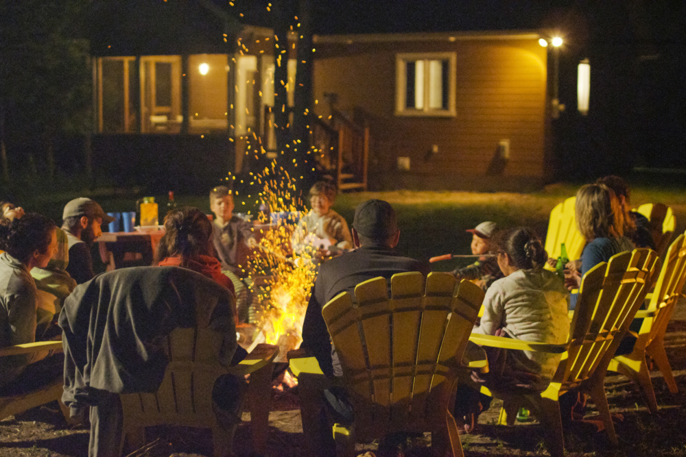 Groupe de personnes réunies autour d’un feu de camp, devant un chalet éclairé, lors d’une soirée en plein air.