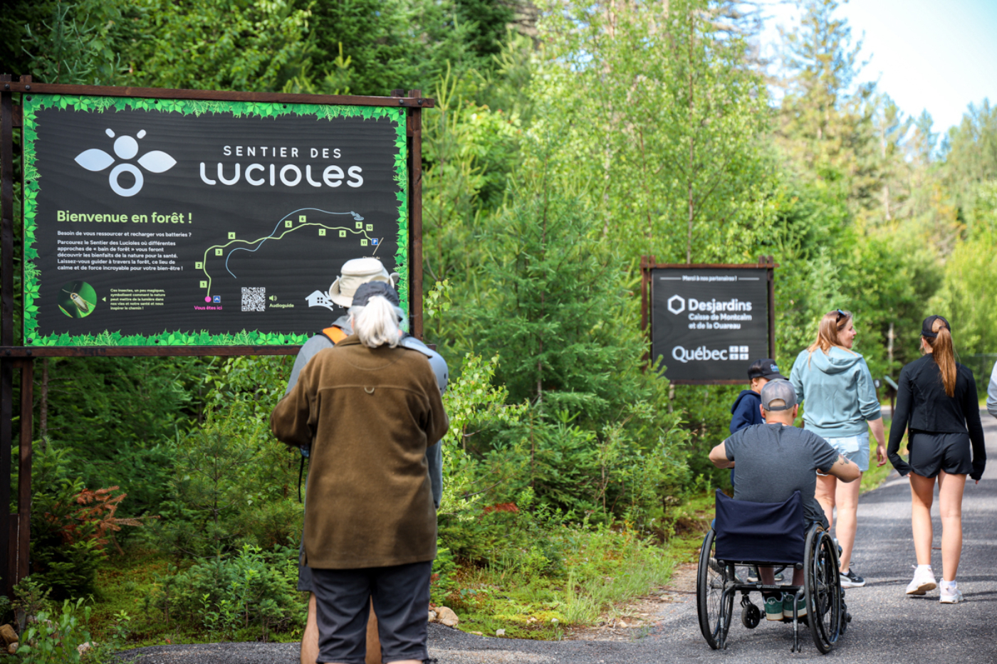 Groupe visitant le Sentier des lucioles en forêt, dont une personne en fauteuil roulant, près d’un panneau de Québec.