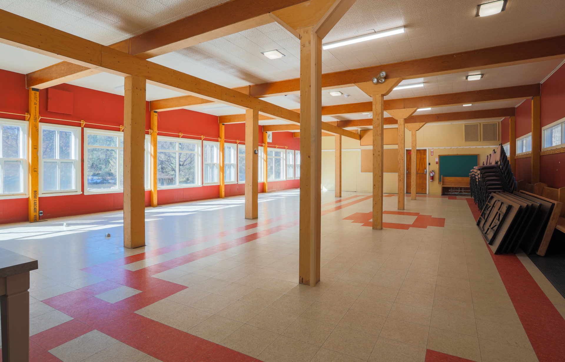 Grande salle polyvalente lumineuse d’un centre de villégiature, avec poutres en bois, fenêtres panoramiques et tables empilées.
