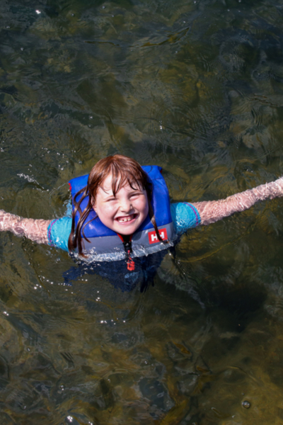 Enfant souriant portant un gilet de sauvetage, nageant dans l’eau près des chalets de Chalets Lanaudière