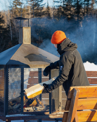 Personne en tuque orange alimentant un feu dans un foyer extérieur enneigé des Chalets Lanaudière.