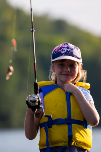 Jeune personne souriante en gilet de sauvetage jaune, tenant une canne à pêche au bord d’un lac près d’un chalet.