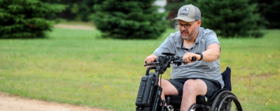 Personne portant une casquette et des lunettes, conduisant un vélo à trois roues adapté sur un sentier, dans un parc verdoyant.