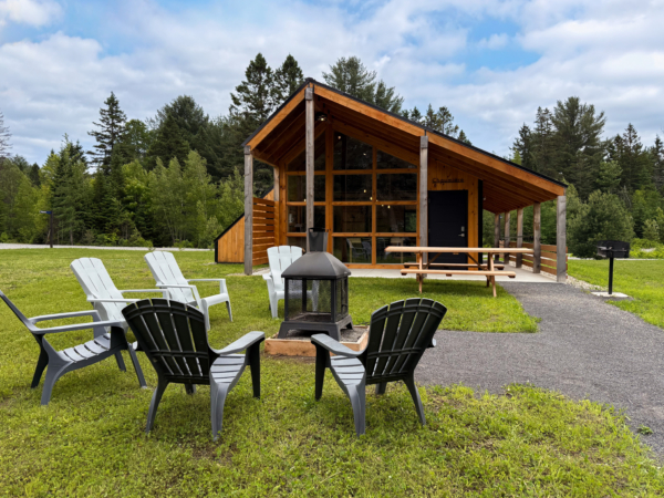 Chalet moderne en bois de Chalets Lanaudière avec terrasse, foyer extérieur et chaises Adirondack dans un cadre forestier.