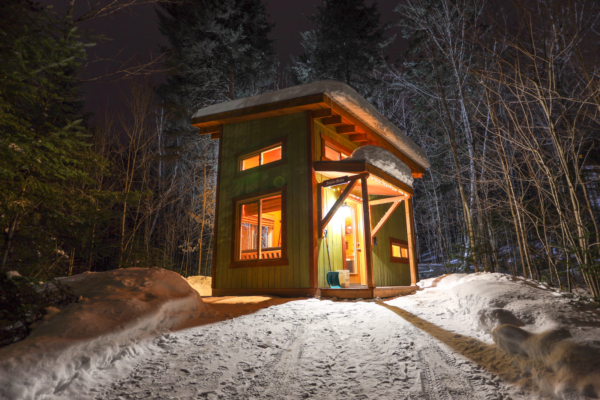 Petit chalet illuminé dans une forêt enneigée, offrant une ambiance chaleureuse pour une escapade chez Chalets Lanaudière.