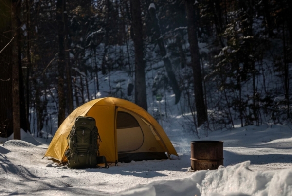 Tente jaune et sac à dos dans un camping enneigé en forêt, avec un foyer métallique à proximité.