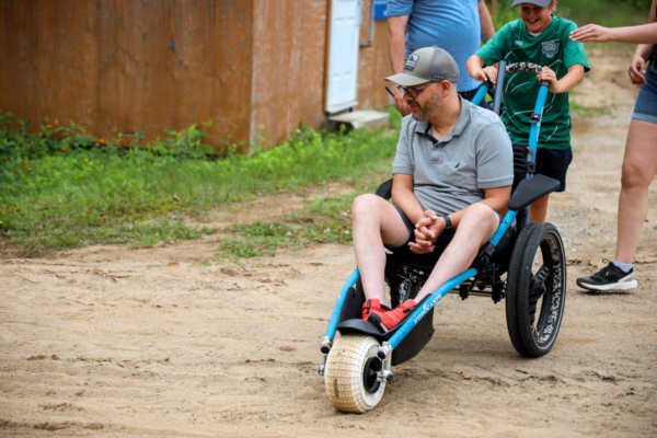 Personne profitant d’une balade en fauteuil tout-terrain, poussée par un enfant sur un sentier sablonneux près d’un chalet.