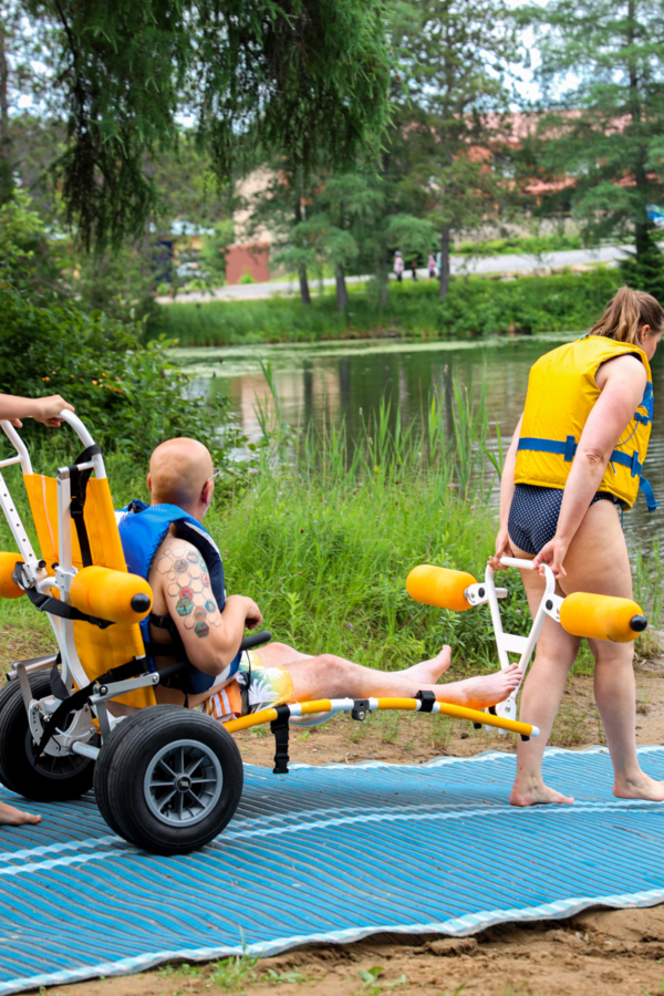 Deux personnes profitent d’un lac près d’un camping, avec un fauteuil roulant amphibie et des gilets de sauvetage.