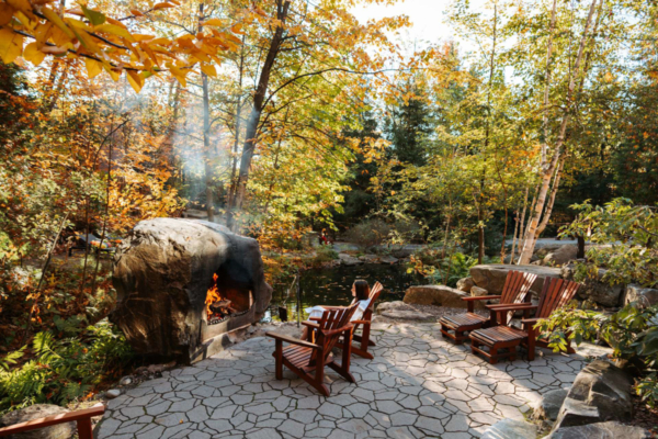 Terrasse en pierre d’un chalet de Chalets Lanaudière, entourée d’arbres, avec foyer extérieur, fauteuils Adirondack et étang.