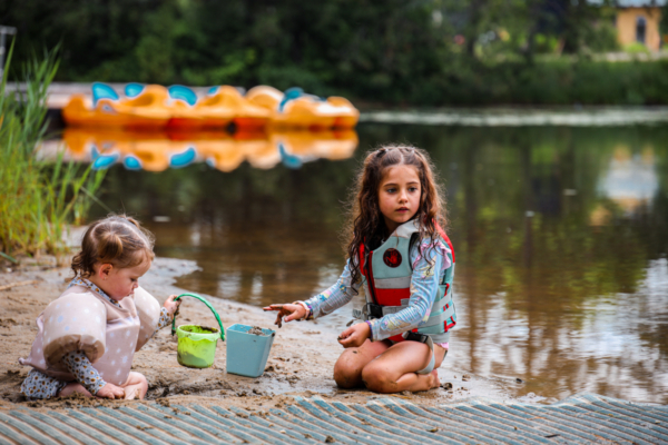 Deux enfants jouent au bord d’un lac près d’un chalet, avec seaux et jouets flottants dans l’eau.
