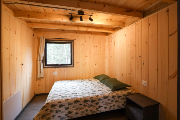 Chambre chaleureuse en bois d’un chalet de Chalets Lanaudière, avec lit double, fenêtre donnant sur la forêt et éclairage au plafond