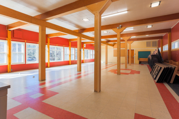 Grande salle polyvalente d’un établissement de Chalets Lanaudière, avec poutres en bois, fenêtres, plancher coloré et chaises empilées.
