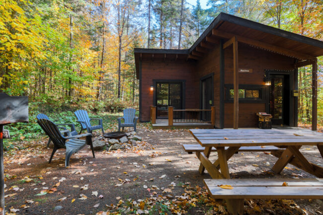 Chalet en bois de Chalets Lanaudière entouré d’arbres colorés, avec terrasse, table à pique-nique, fauteuils et foyer extérieur.