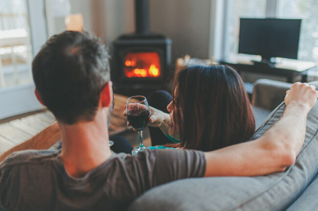 Couple profitant d’un moment chaleureux dans un chalet, un verre de vin à la main près du poêle à bois.