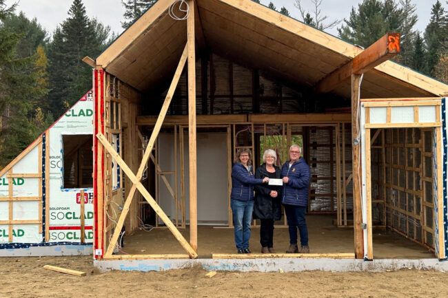 Trois personnes devant un chalet en construction, entourées de structures en bois et de matériaux isolants, dans un cadre forestier.