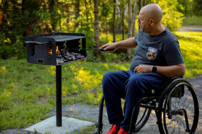 Personne en fauteuil roulant faisant griller des aliments sur un barbecue extérieur, dans un décor boisé de Chalets Lanaudière.