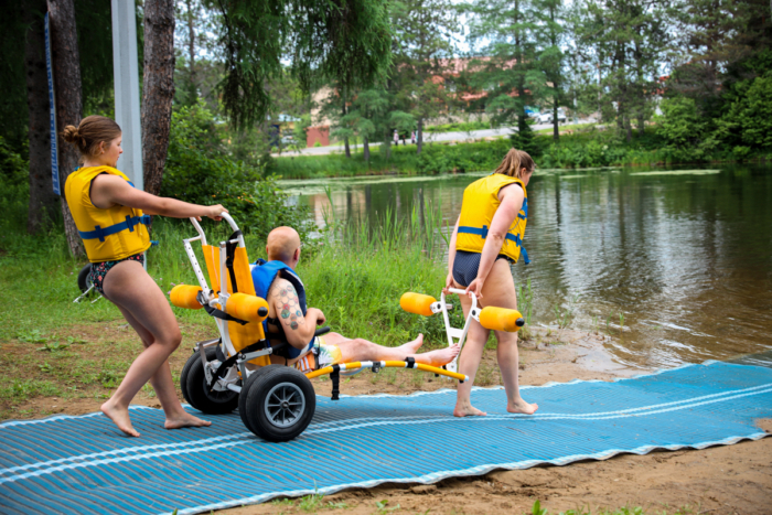 Des vacanciers accompagnent une personne en fauteuil amphibie vers le lac, dans un camping de Chalets Lanaudière.