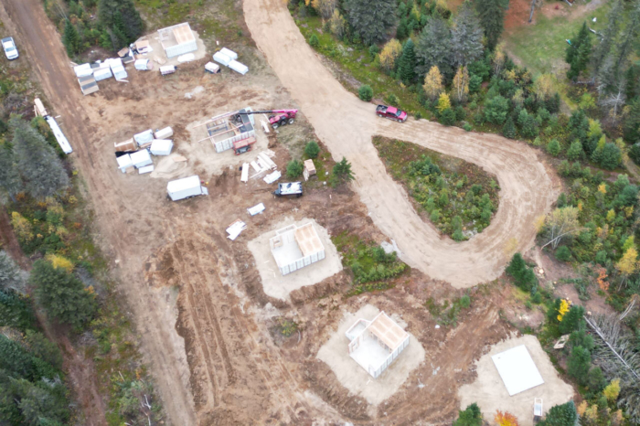 Vue aérienne d’un chantier de construction de chalets en forêt, avec fondations, matériaux et chemin de terre sinueux.