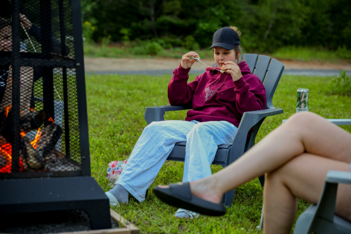Personne assise près d’un feu de camp, faisant griller une guimauve, tandis qu’une autre se détend à proximité dans un camping.