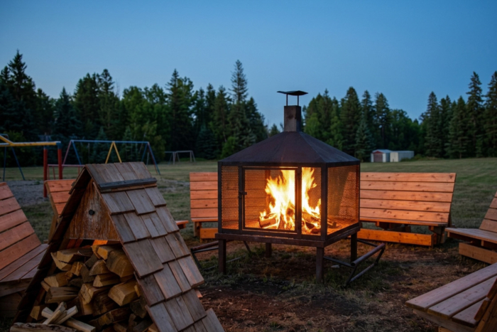 Foyer extérieur dans un abri grillagé, entouré de bancs, de bois de chauffage et d’une aire de jeux au camping Chalets Lanaudière.
