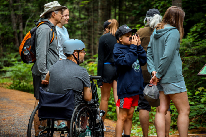 Groupe profitant d’une randonnée en forêt près de leur hébergement aux Chalets Lanaudière, avec une personne en fauteuil roulant et un enfant aux jumelles.