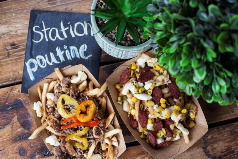 Deux portions de poutine garnies de fromage, légumes et viande sur une table en bois, avec une pancarte « Station poutine ».
