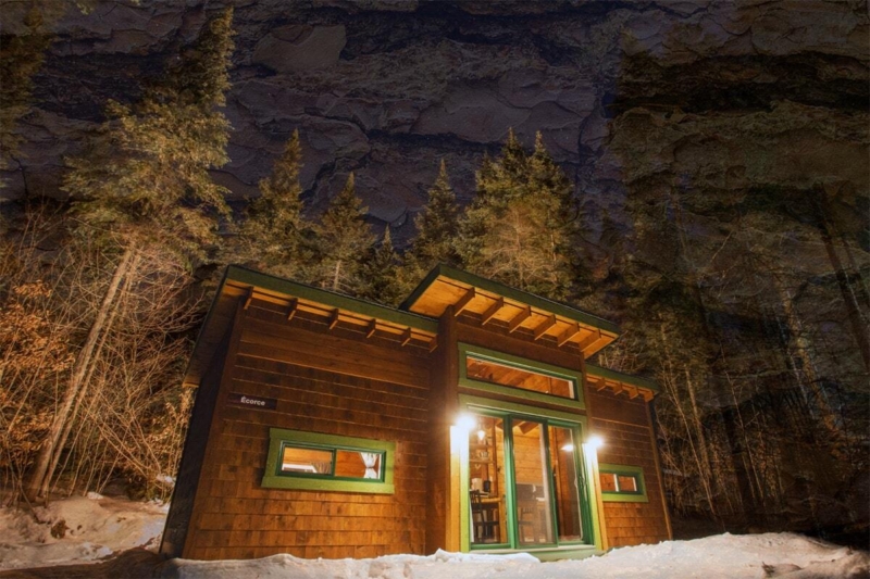 Chalet en bois chaleureux de Chalets Lanaudière, éclairé le soir et entouré de forêt enneigée au pied d’une falaise rocheuse.