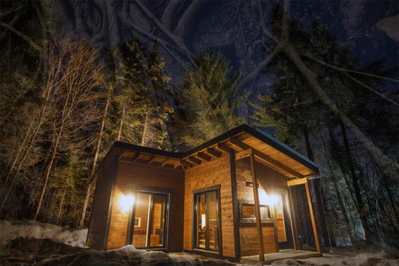 Chalet chaleureux illuminé la nuit, entouré de forêt et de neige, offrant une ambiance paisible chez Chalets Lanaudière.