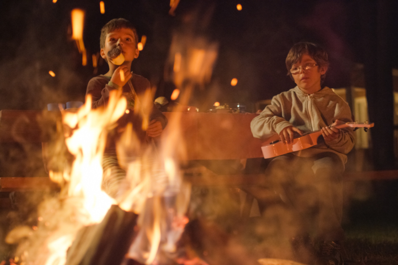 Deux enfants profitent d’une soirée de camping à Chalets Lanaudière autour d’un feu de camp, l’un chantant et l’autre jouant du ukulélé.