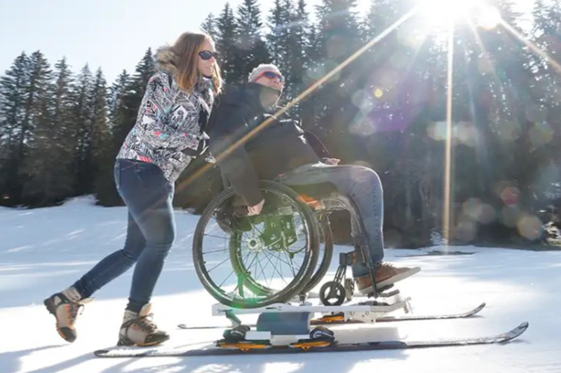 Personne poussant une personne en fauteuil adapté au ski sur une piste enneigée près des chalets de Lanaudière.
