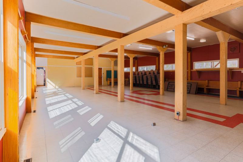 Grande salle commune d’un chalet, avec poutres en bois, tables et chaises, baignée de lumière naturelle.