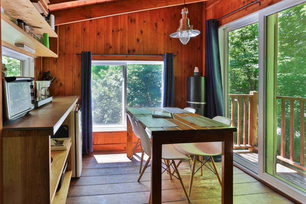 Intérieur chaleureux d’une cabane avec cuisinette, coin repas et grandes fenêtres donnant sur une terrasse entourée d’arbres.