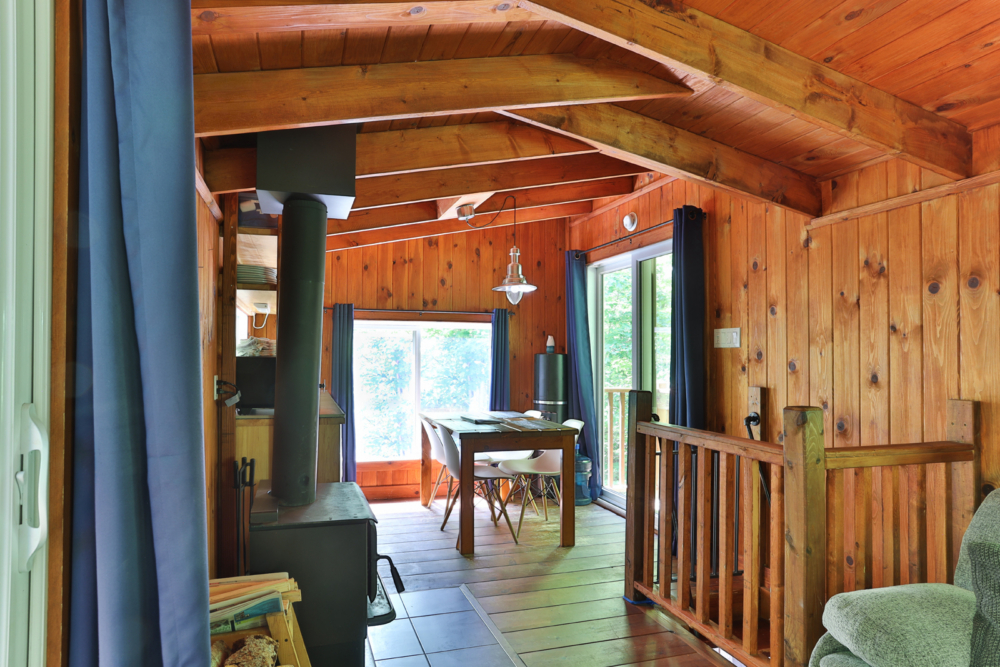 Intérieur chaleureux d’une cabane en bois, avec poêle, salle à manger et grandes fenêtres donnant sur la nature.