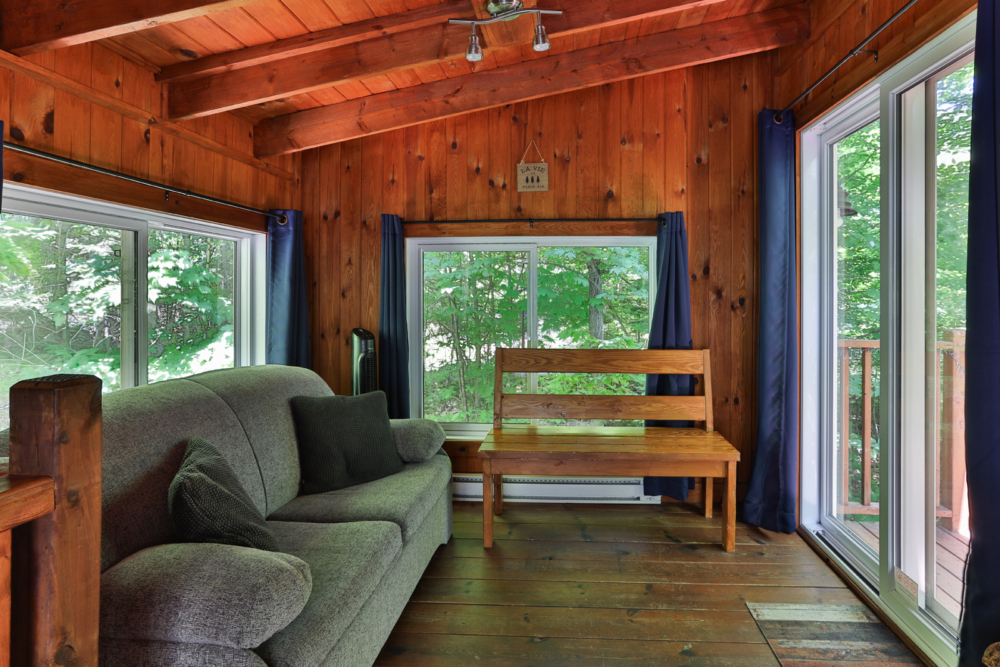 Salon chaleureux d’une cabane avec canapé, banc en bois et grandes fenêtres donnant sur la forêt.