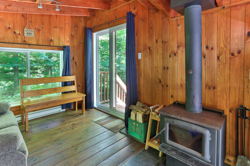 Intérieur chaleureux d’une cabane en bois avec poêle à bois, banc, fenêtres et porte donnant sur la forêt.