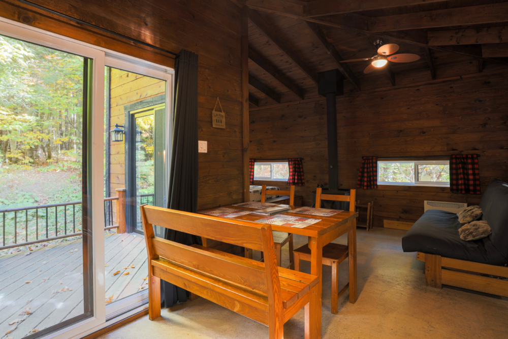 Intérieur chaleureux d’une cabane avec table à manger en bois, poêle à bois et accès à la terrasse.