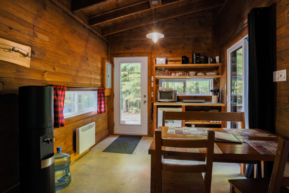 Intérieur chaleureux d’une cabane, avec coin repas, cuisinette équipée et vue sur la forêt.