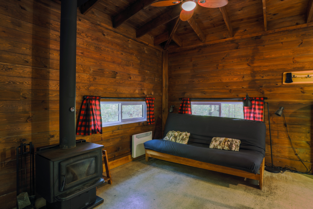 Intérieur chaleureux d’une cabane avec poêle à bois, divan-lit et rideaux à carreaux rouges.