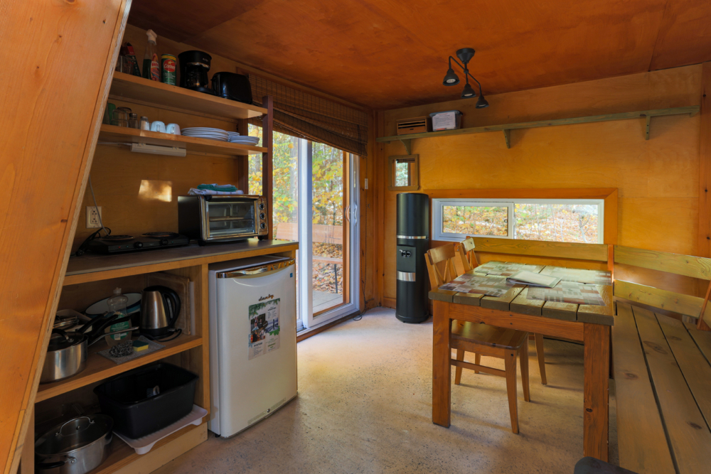 Intérieur chaleureux d’une cabane avec cuisinette tout équipée, mini-réfrigérateur, coin repas et vue sur la forêt.