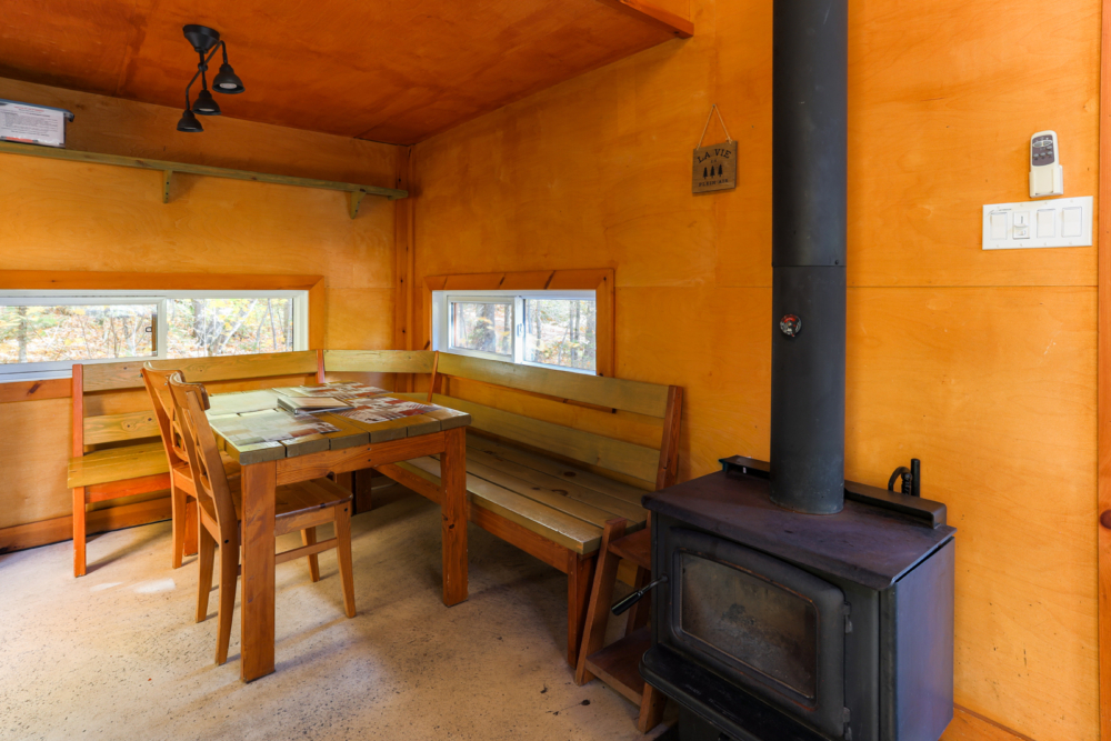 Intérieur chaleureux d’une cabane avec table à manger, banquettes en bois et poêle à bois.