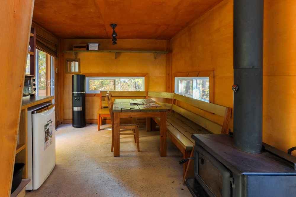Intérieur chaleureux d’une cabane avec table à manger, banquettes en bois et poêle à bois.