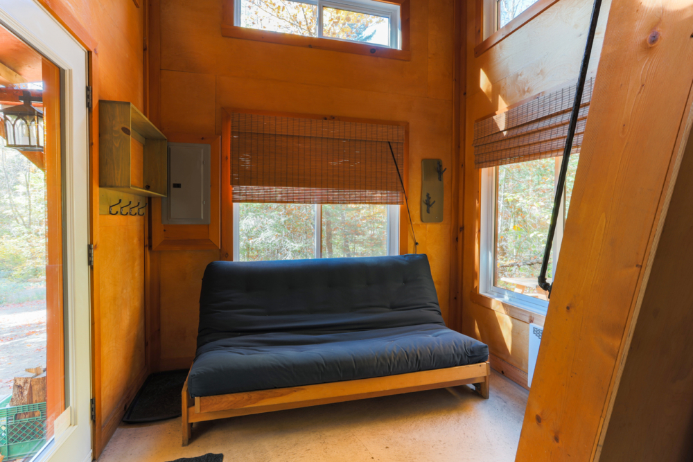 Intérieur chaleureux d’une cabane avec futon bleu, grandes fenêtres et vue sur la forêt.