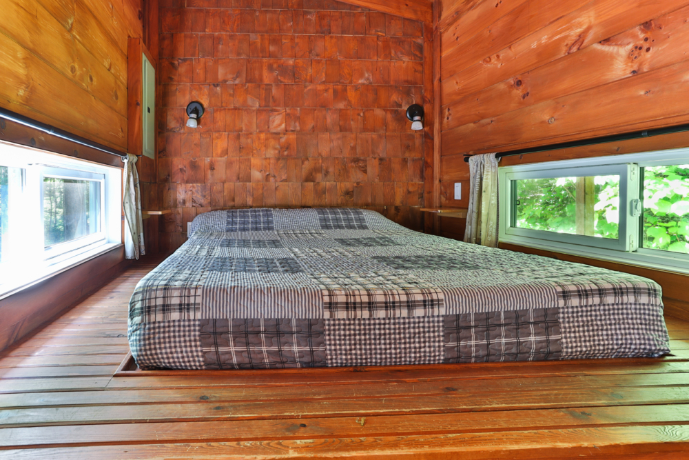 Chambre chaleureuse d’une cabane avec lit double, murs en bois et fenêtres donnant sur la forêt.