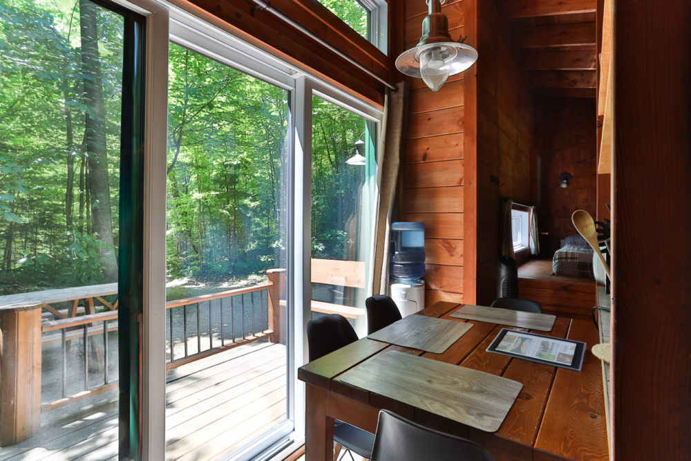 Coin repas d’une cabane en bois avec grande fenêtre donnant sur la forêt et la terrasse.