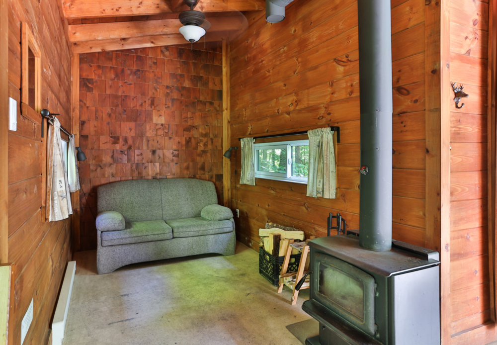 Intérieur chaleureux d’une cabane en bois avec divan, poêle à bois, fenêtre et bûches.
