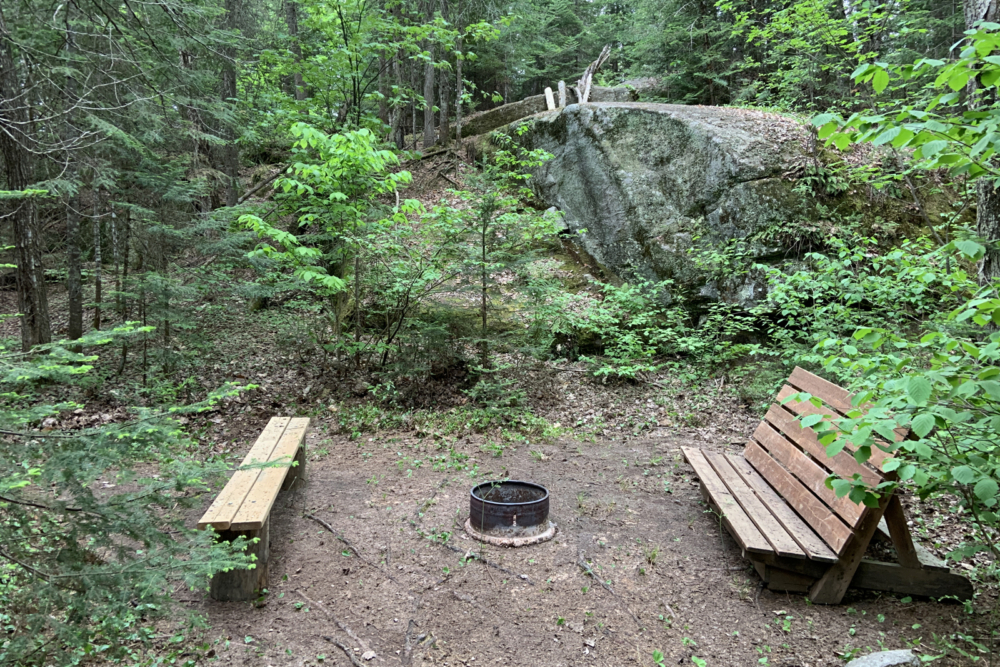 Site de camping avec espace de feu de camp en forêt, avec bancs en bois et grand rocher.