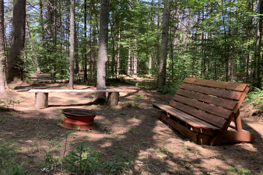Wooded campsite with a rustic bench, fire pit and picnic table, offering a peaceful outdoor amenity at Chalets Lanaudière.